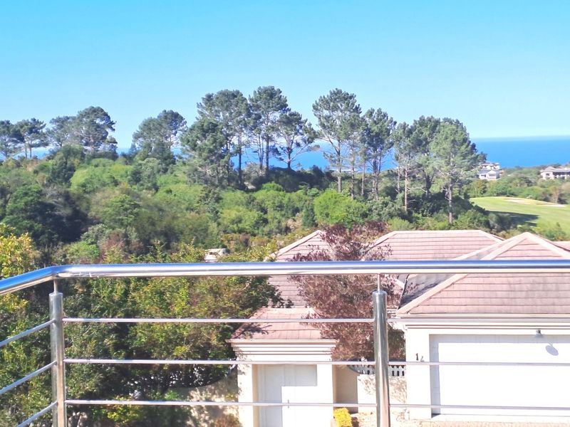 Elevated view overlooking a residential neighborhood with lush greenery, mature trees, and a distant ocean horizon, framed by a modern stainless steel balustrade.