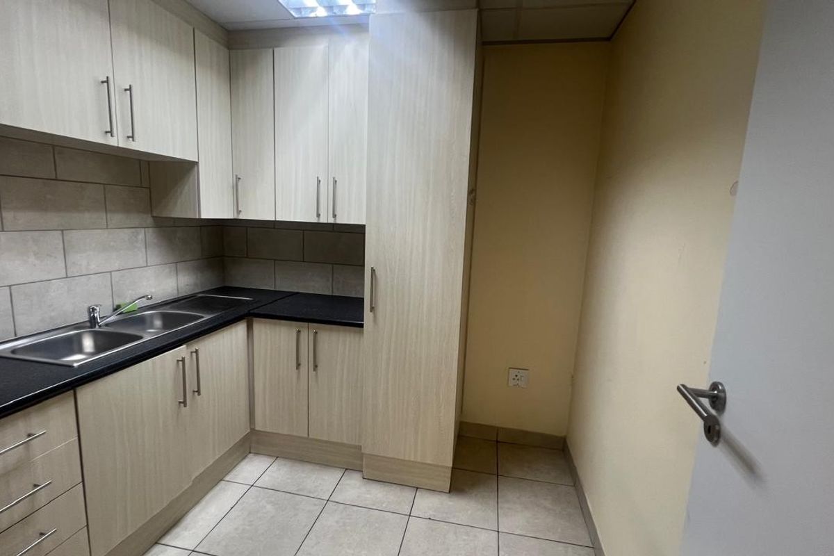 A compact, functional kitchen area featuring light wood-finish cabinetry, dark laminate countertops, a stainless steel double sink, and neutral tiled flooring.