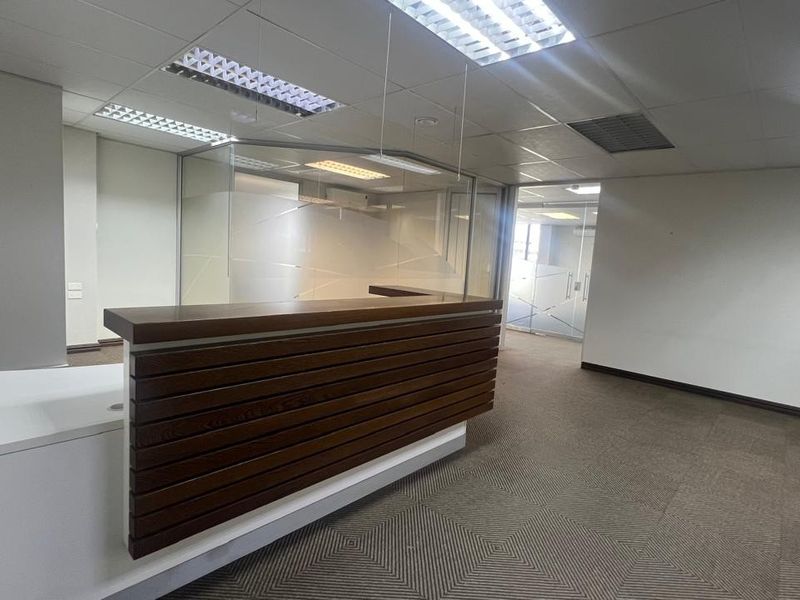 A professional commercial reception area featuring a built-in wooden reception desk, glass partitions, suspended ceilings, and neutral carpet flooring.
