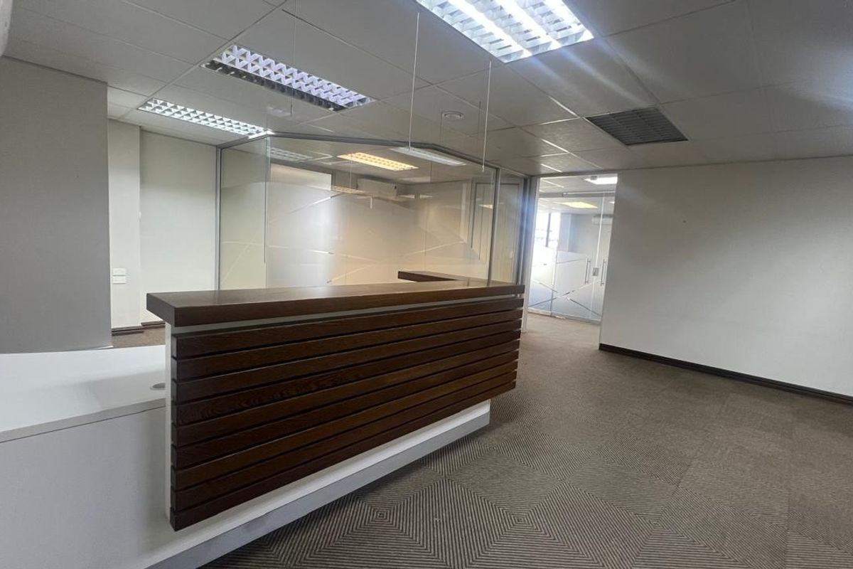 A professional commercial reception area featuring a built-in wooden reception desk, glass partitions, suspended ceilings, and neutral carpet flooring.