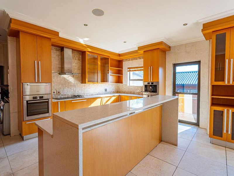 A spacious kitchen featuring light wood cabinetry, stone countertops, built-in appliances including an oven and coffee machine, and tiled flooring with direct outdoor access.