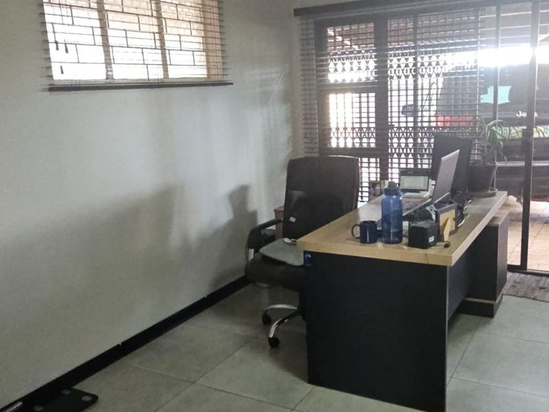 A functional home office space featuring tiled flooring, large windows with blinds, and direct access to the driveway area.