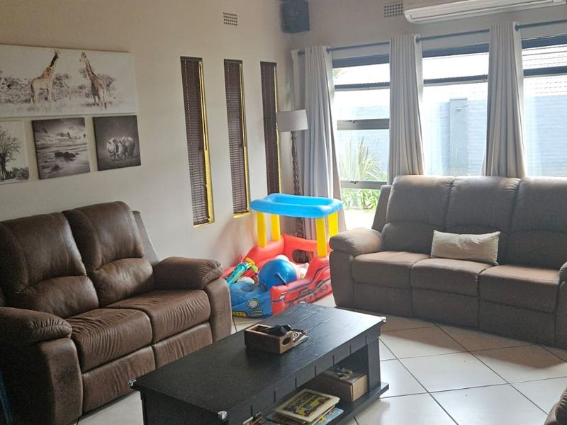 A comfortable living room featuring large aluminium-framed windows, tiled flooring, and a wall-mounted air conditioning unit, offering views of the rear garden.