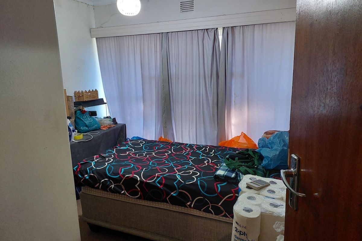 A bedroom featuring a large window with curtain rails and a ceiling vent, showing signs of wear including cracked plaster and peeling paint.