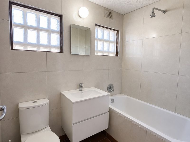 A functional bathroom featuring a wall-mounted vanity, bathtub with shower head, and glass block windows, finished with neutral wall tiles and terracotta floor tiles.