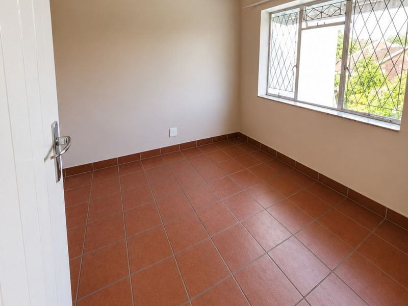A bright, empty bedroom featuring terracotta-style floor tiles, an exposed ceiling beam, and large windows with security bars.
