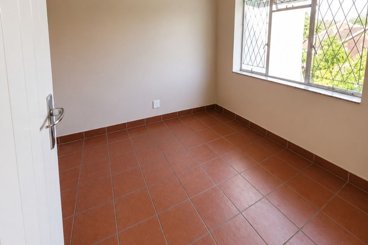 A bright, empty bedroom featuring terracotta-style floor tiles, an exposed ceiling beam, and large windows with security bars.