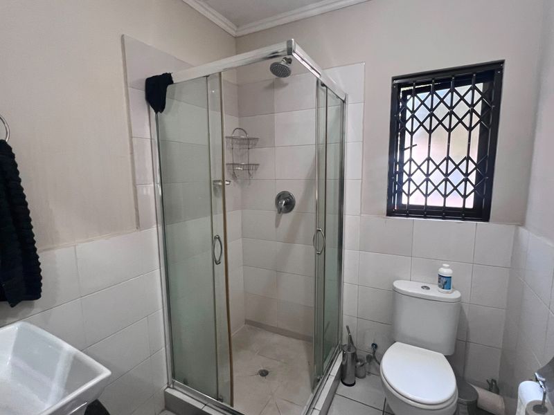 A functional bathroom featuring a glass-enclosed shower, white wall tiling, and a standard toilet with a security-gated window.