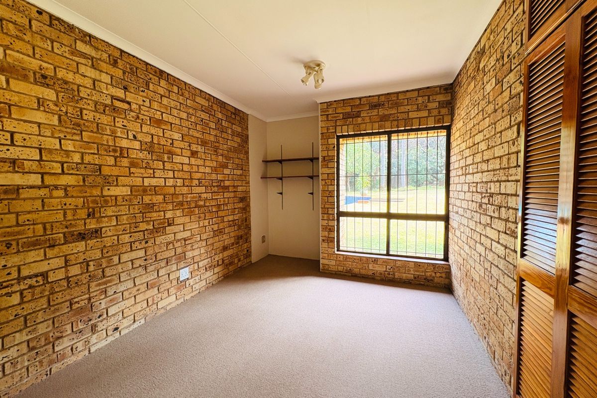 A bright, empty bedroom featuring exposed brick walls, built-in wooden louvered wardrobes, and a large window with security bars overlooking the garden.