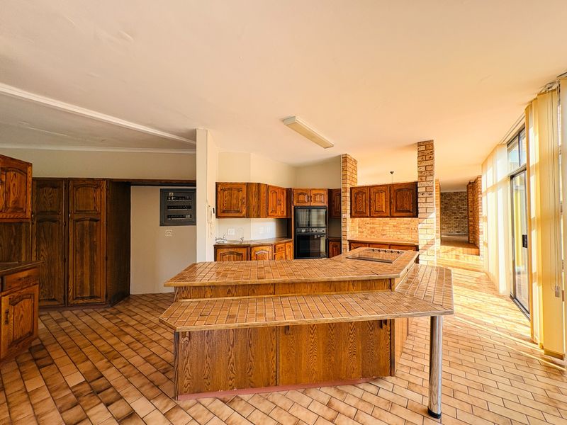 Spacious kitchen featuring a large tiled island, built-in oven and hob, wooden cabinetry, and exposed brick accents, offering significant potential for modernization.