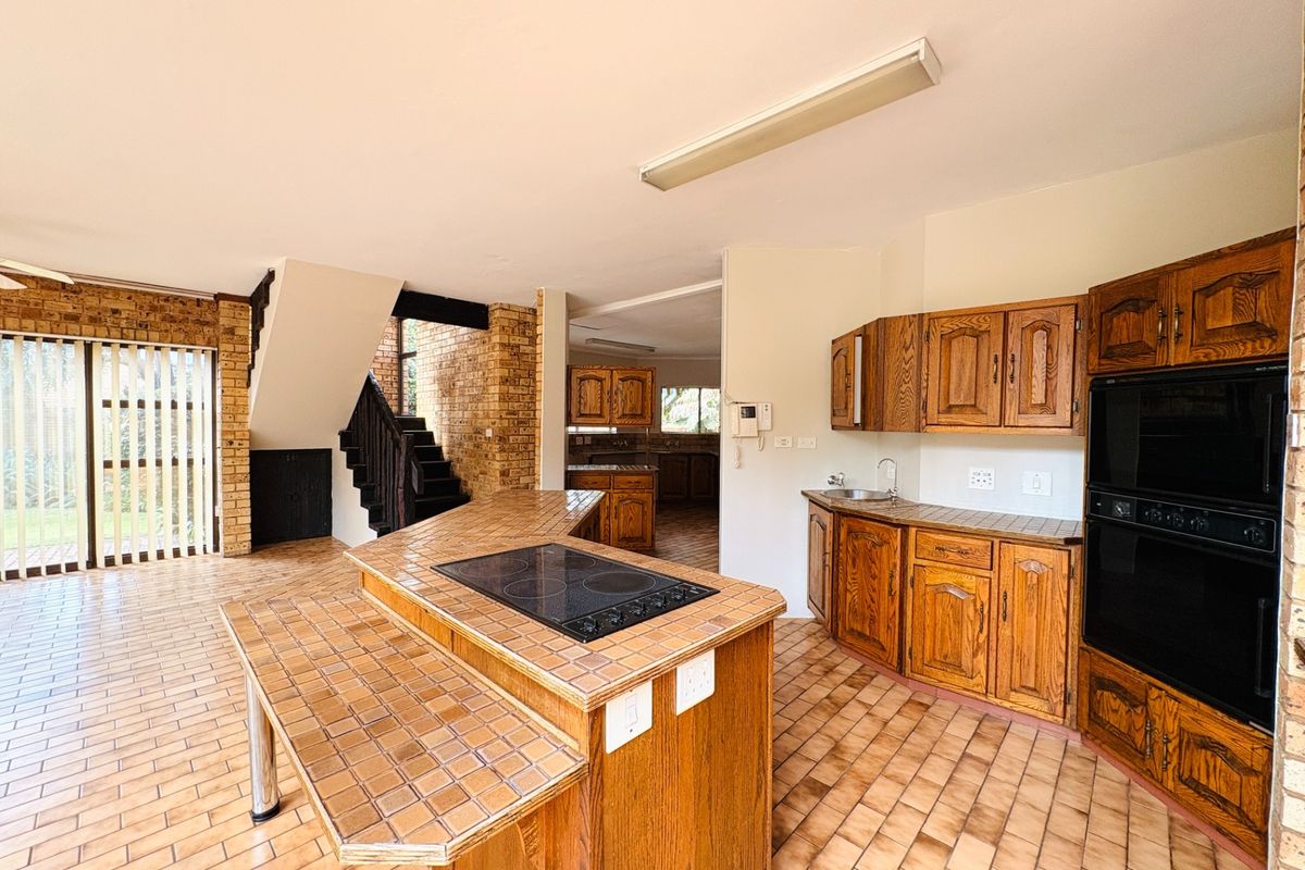 Spacious kitchen featuring a central island with built-in hob, double oven, wooden cabinetry, and tiled flooring, with an adjacent staircase and exposed brick accents.