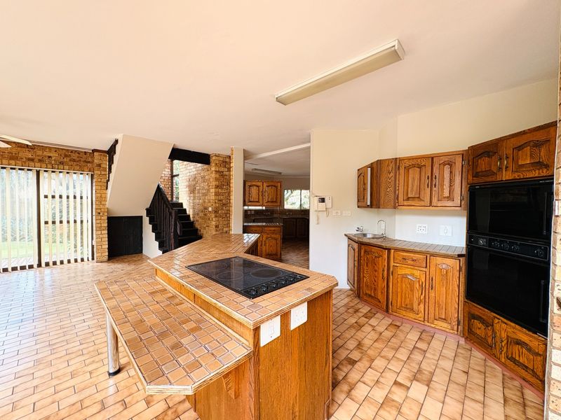Spacious kitchen featuring a central island with built-in hob, double oven, wooden cabinetry, and tiled flooring, with an adjacent staircase and exposed brick accents.