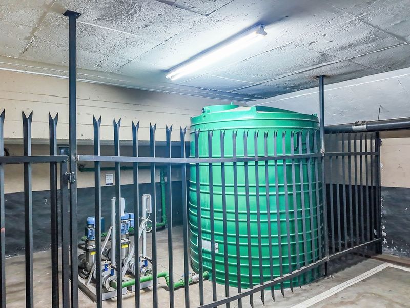 Utility area featuring a large green water storage tank and associated pump system, secured within a fenced enclosure with exposed concrete ceiling.