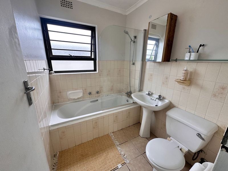 A functional bathroom featuring a bathtub with shower attachment, pedestal basin, and toilet, finished with neutral wall and floor tiling.