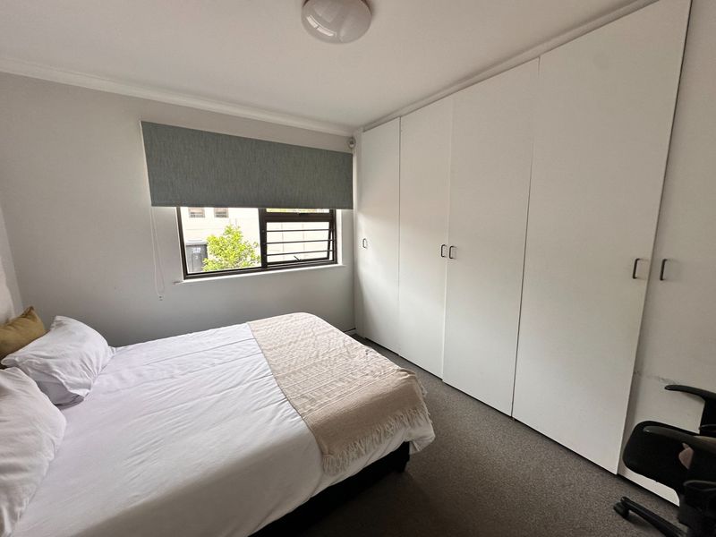 A well-maintained bedroom featuring neutral walls, carpeted flooring, and extensive built-in cupboard storage with a window fitted with a roller blind.