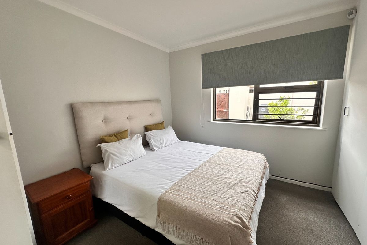 A well-maintained bedroom featuring neutral wall finishes, carpeted flooring, a built-in cupboard, and a window fitted with security bars and a roller blind.