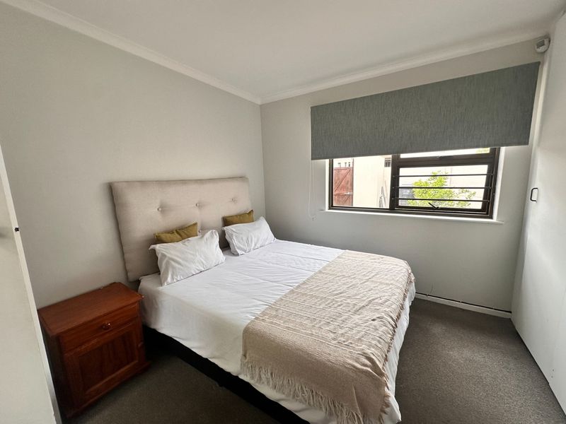 A well-maintained bedroom featuring neutral wall finishes, carpeted flooring, a built-in cupboard, and a window fitted with security bars and a roller blind.