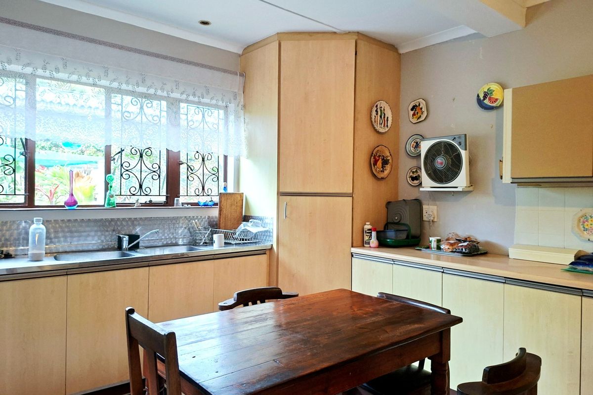 A functional kitchen featuring light wood-tone cabinetry, laminate countertops, a stainless steel double sink, and a tiled splashback with security-barred windows.