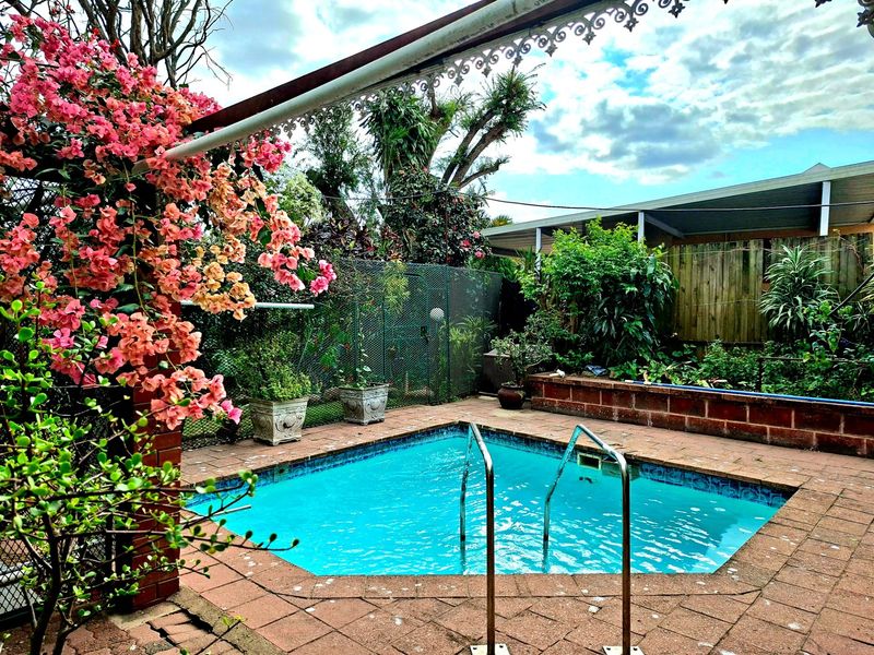 Outdoor pool area featuring a paved surround, brick retaining wall, and metal pergola structure, complemented by established garden landscaping.