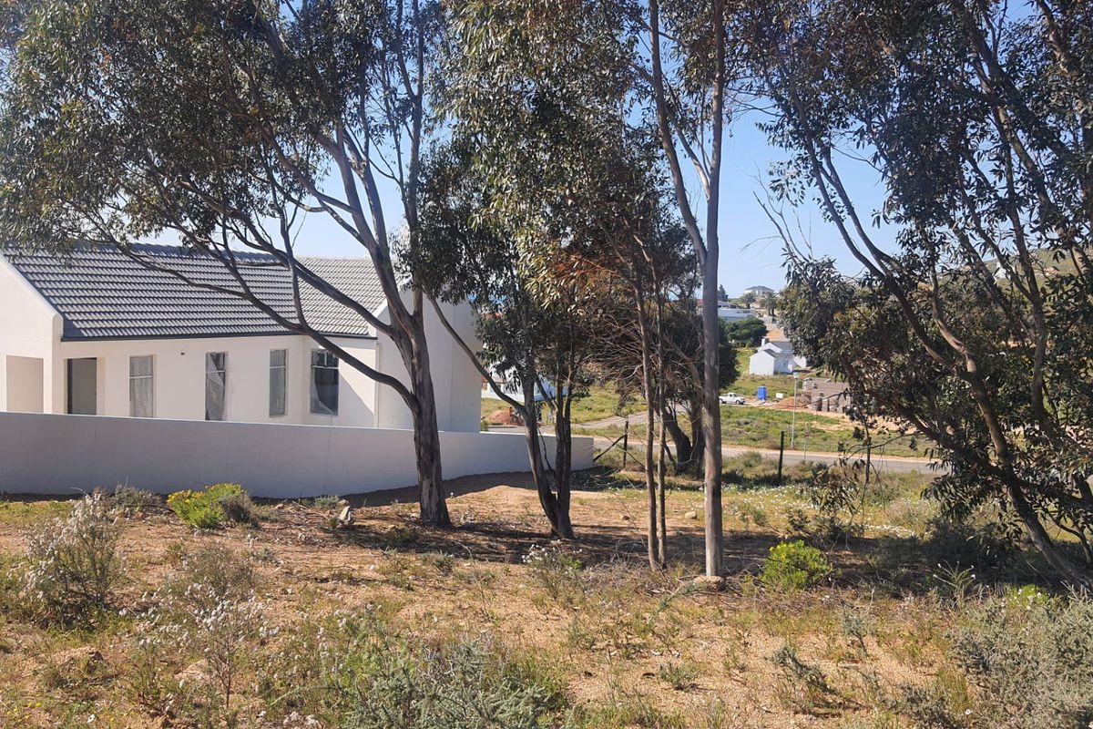 A view of a residential property featuring a white exterior facade, tiled roof, and boundary wall, set within a natural landscape with mature trees.