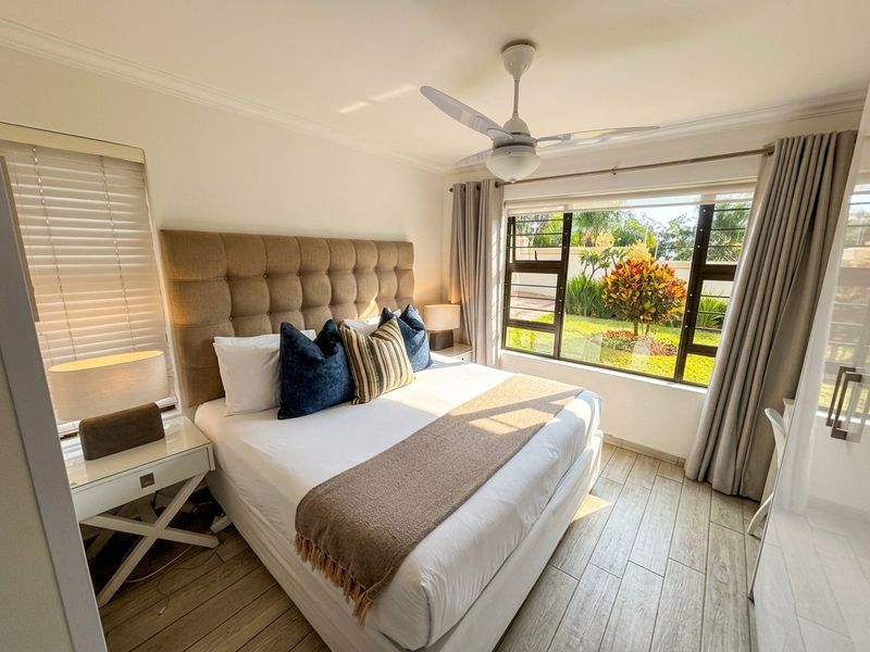 A bright and well-maintained bedroom featuring tiled flooring, a ceiling fan, and large windows with views of the garden.