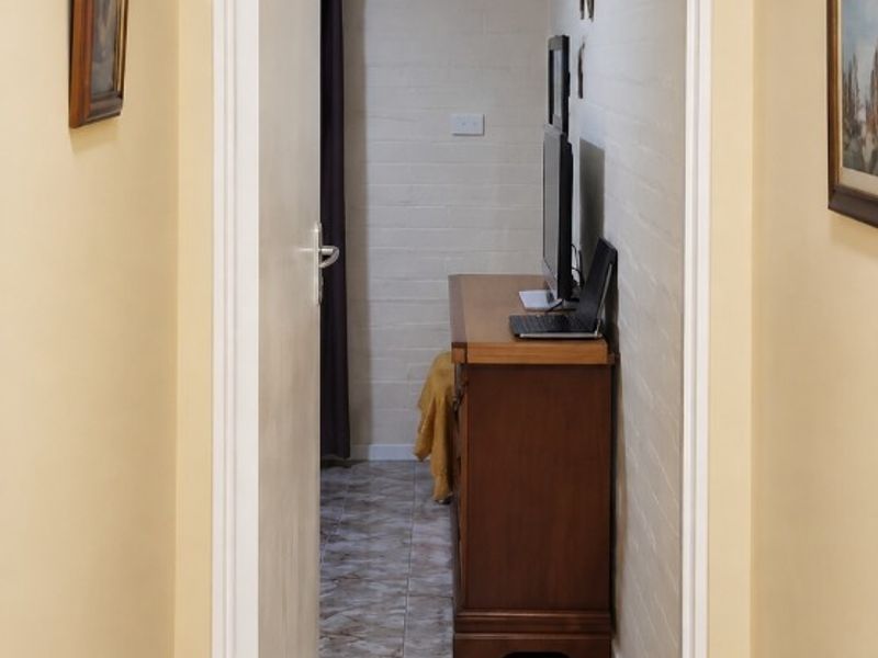 A narrow hallway featuring tiled flooring and painted brick walls, leading into an adjacent room with a ceiling access hatch.