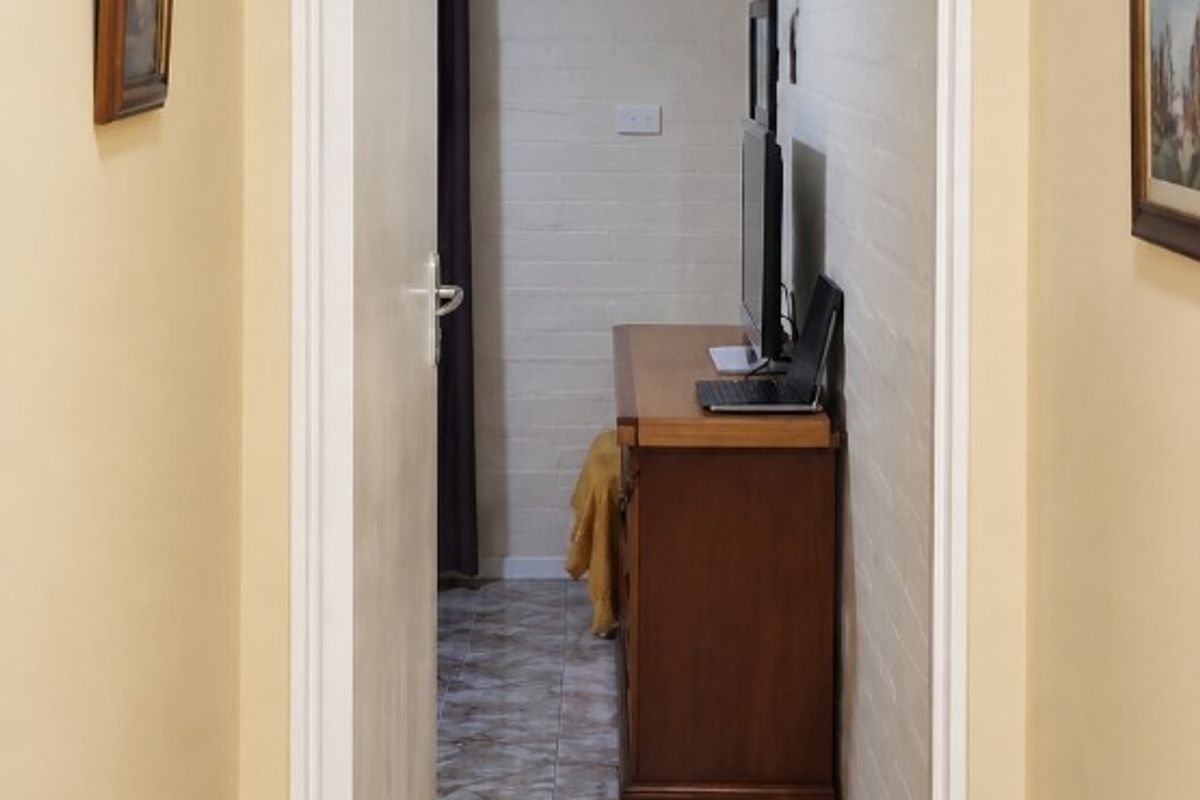 A narrow hallway featuring tiled flooring and painted brick walls, leading into an adjacent room with a ceiling access hatch.