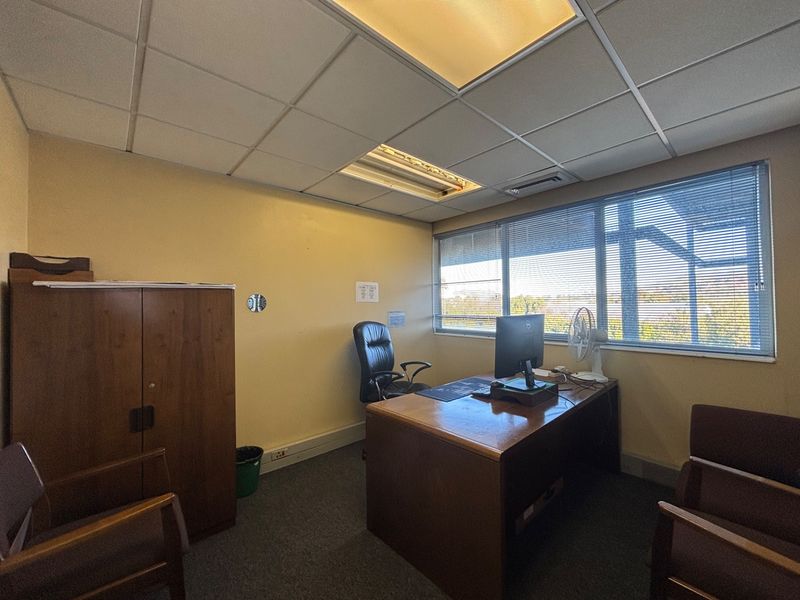 A functional office space featuring carpeted flooring, a suspended ceiling with integrated lighting, and large windows with venetian blinds providing natural light.