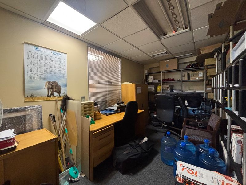 A functional office space featuring a suspended ceiling with recessed lighting, carpeted flooring, and venetian blinds, currently used for storage and workspace.