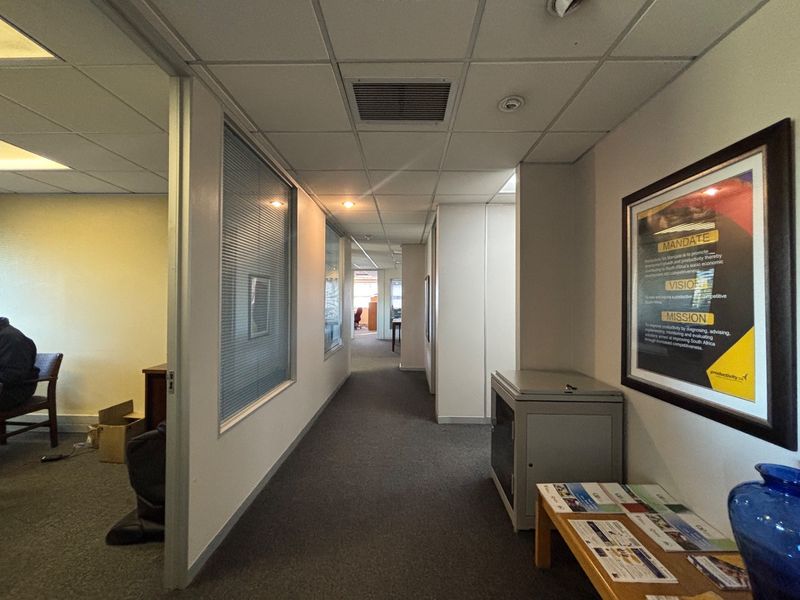 A functional office hallway featuring carpeted flooring, suspended ceilings with recessed lighting, and internal glass partitions providing views into adjacent office spaces.