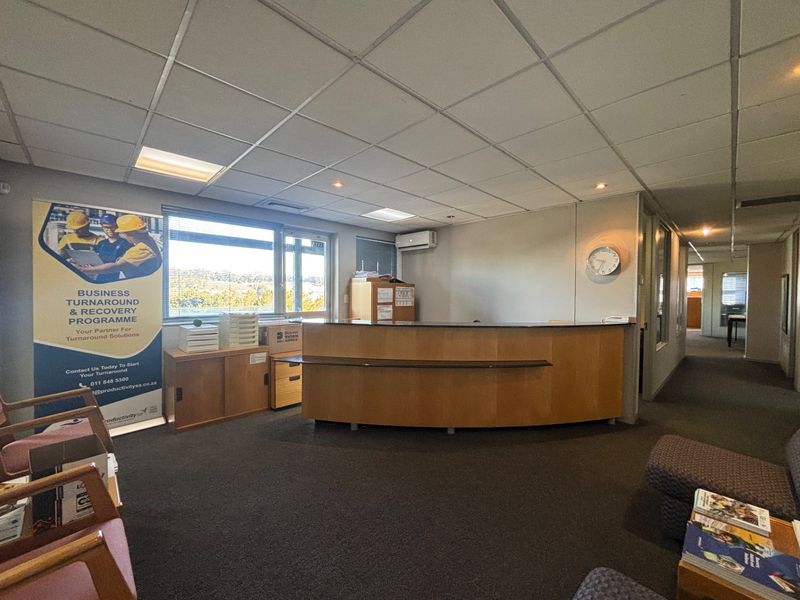 A professional office reception area featuring a built-in curved reception desk, suspended ceiling tiles, and carpeted flooring with natural light from the window.