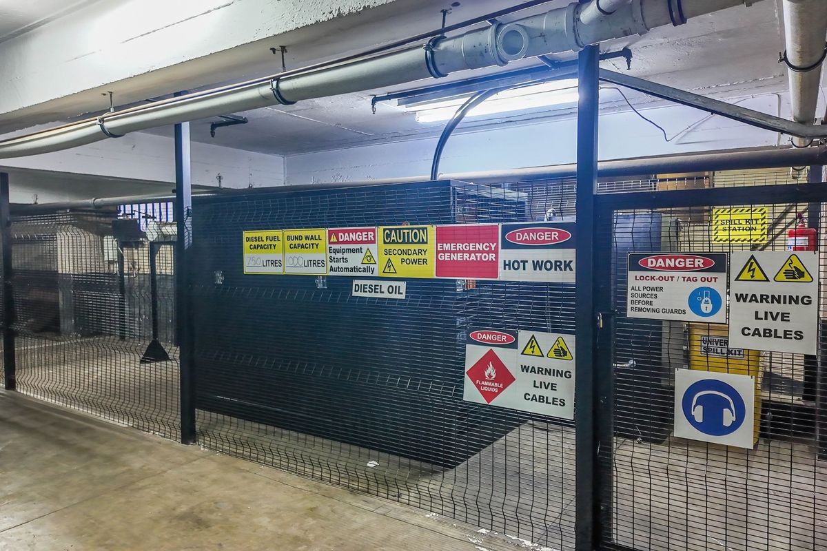 A secure utility area housing a diesel generator, featuring industrial metal fencing, exposed overhead piping, and comprehensive safety signage.