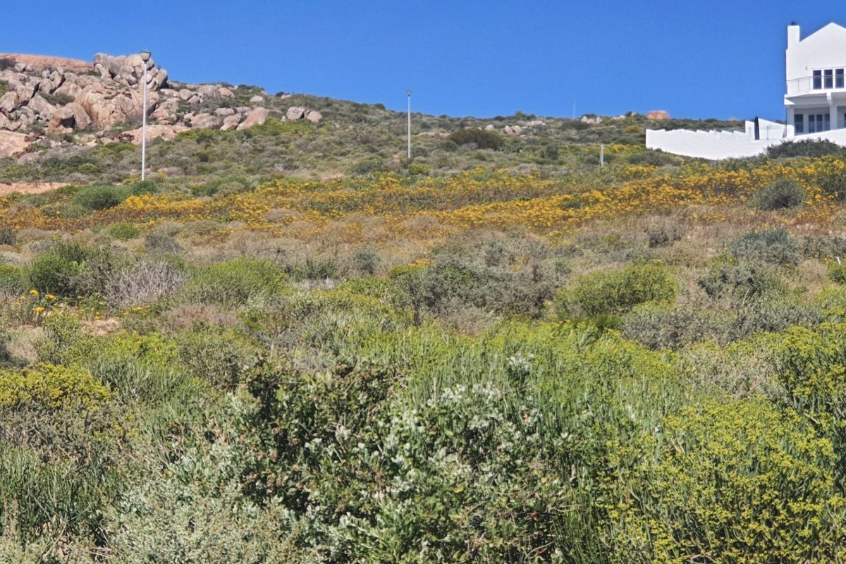 A scenic view of a sloped residential plot featuring natural coastal vegetation, rocky outcrops, and a neighboring property in the background.