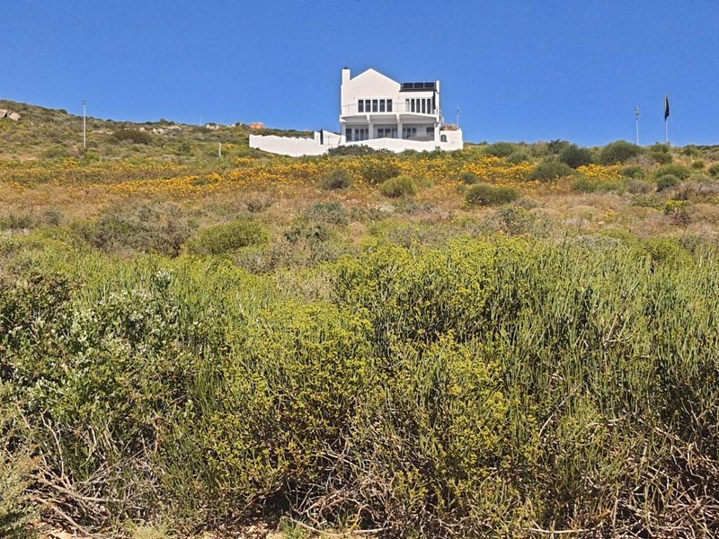A view of a residential property situated on a sloped, natural landscape, featuring a white exterior, balcony, and roof-mounted solar panels.