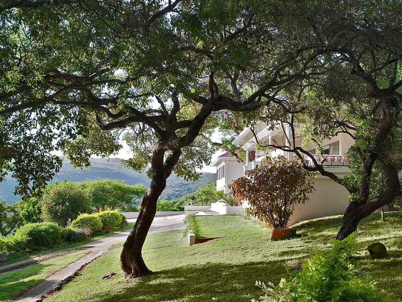 A scenic view of the property's rear exterior, featuring a landscaped garden, mature trees, a paved driveway, and a balcony overlooking the surrounding landscape.