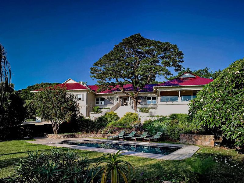 A rear view of the farmhouse featuring a swimming pool, landscaped garden, stone retaining walls, and a covered patio area under a red metal roof.