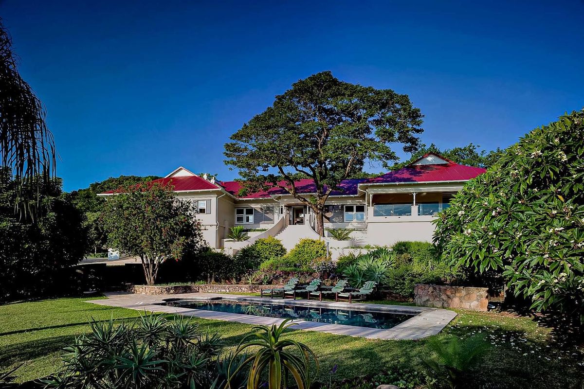 A rear view of the farmhouse featuring a swimming pool, landscaped garden, stone retaining walls, and a covered patio area under a red metal roof.