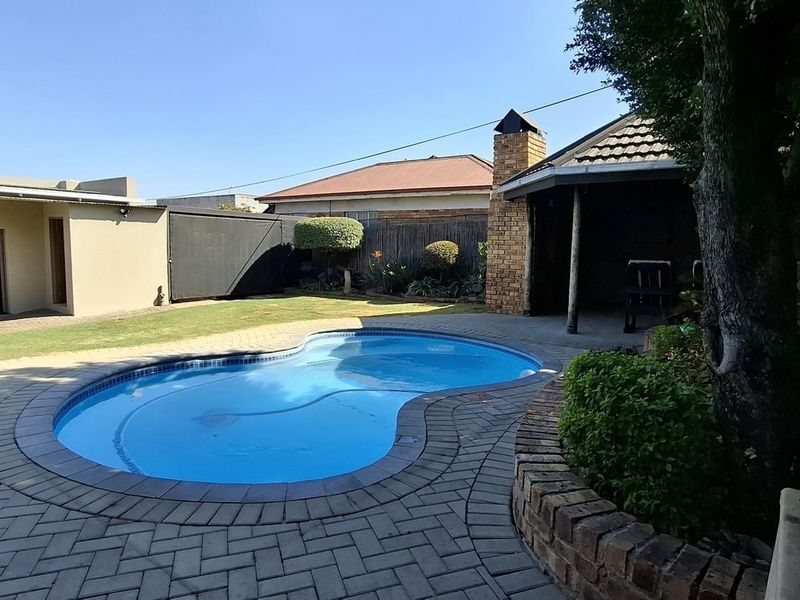 A well-maintained backyard featuring a kidney-shaped swimming pool, brick paved surround, and a covered outdoor entertainment area with a built-in brick fireplace.