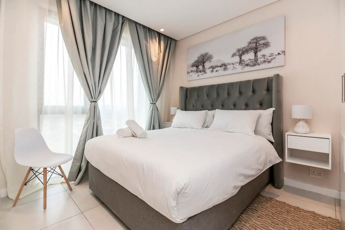 A well-maintained bedroom featuring neutral tones, tiled flooring, recessed lighting, and large windows with elegant floor-to-ceiling curtains.