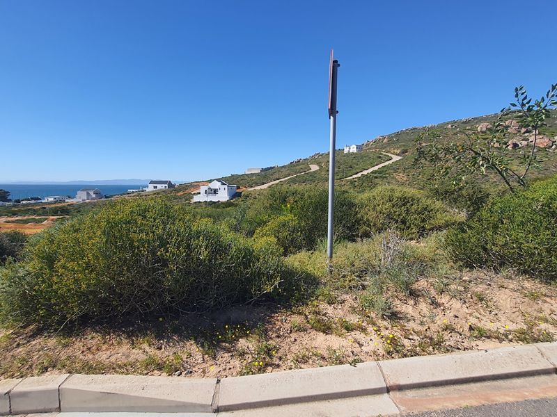 A view of a vacant residential plot featuring natural coastal vegetation, concrete kerbing, and distant ocean views in a developing estate.