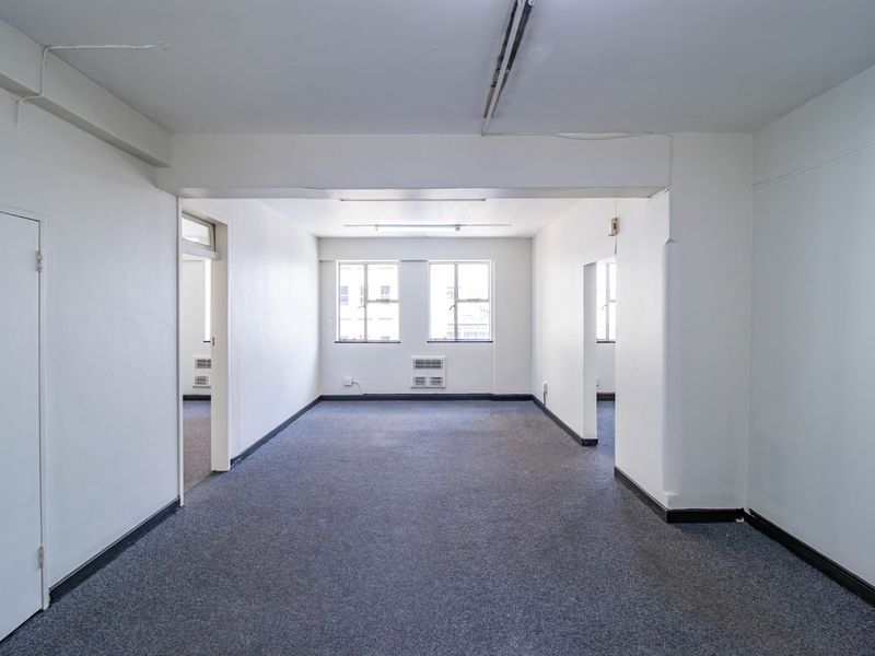 An empty, open-plan commercial office space featuring grey carpet flooring, window-mounted air conditioning, and basic fluorescent lighting.