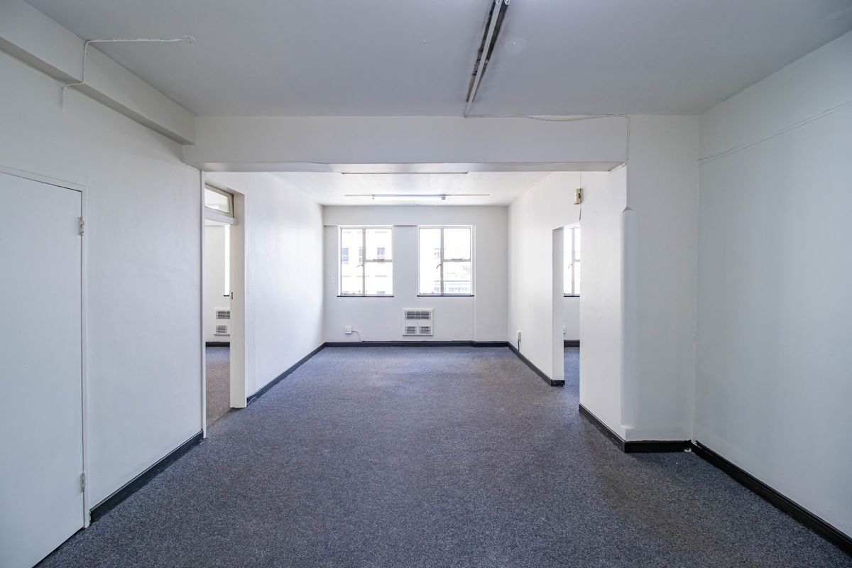 An empty, open-plan commercial office space featuring grey carpet flooring, window-mounted air conditioning, and basic fluorescent lighting.