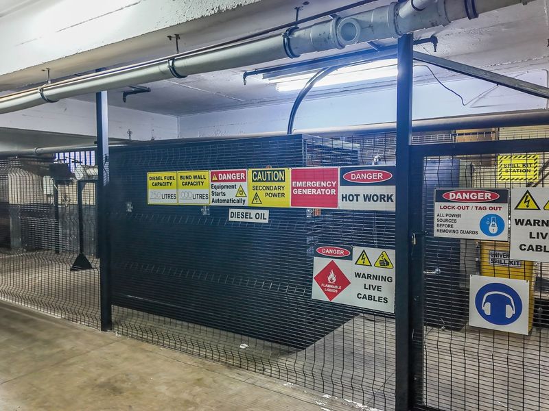 A secure utility area housing a diesel generator, featuring industrial metal fencing, exposed overhead piping, and comprehensive safety signage.