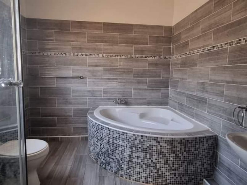 A modern bathroom featuring a corner bathtub with mosaic tiling, a separate glass shower enclosure, a pedestal basin, and neutral grey wall tiles.