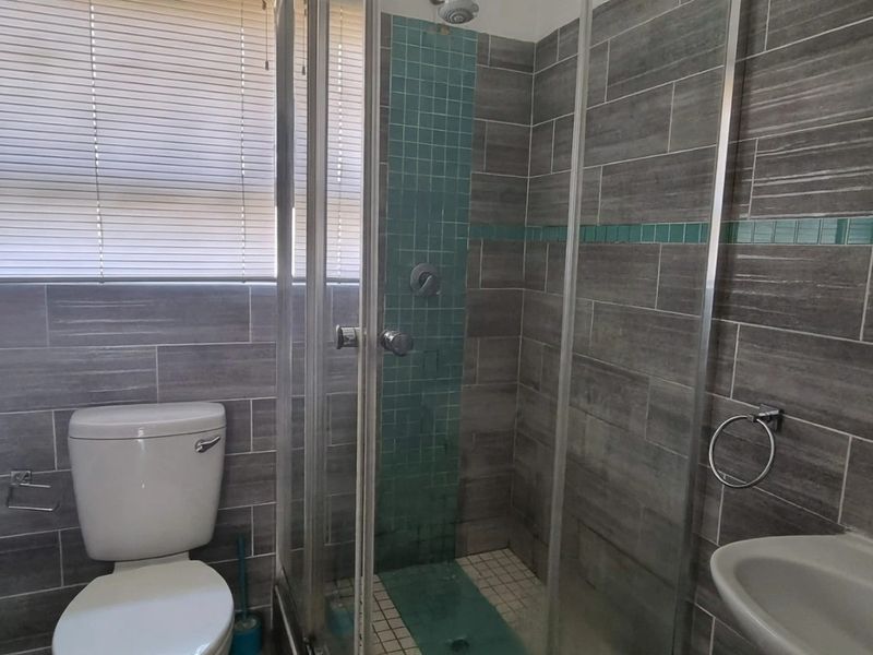 A functional bathroom featuring a glass shower enclosure, bathtub, pedestal basin, and grey tiled walls with teal accent detailing.