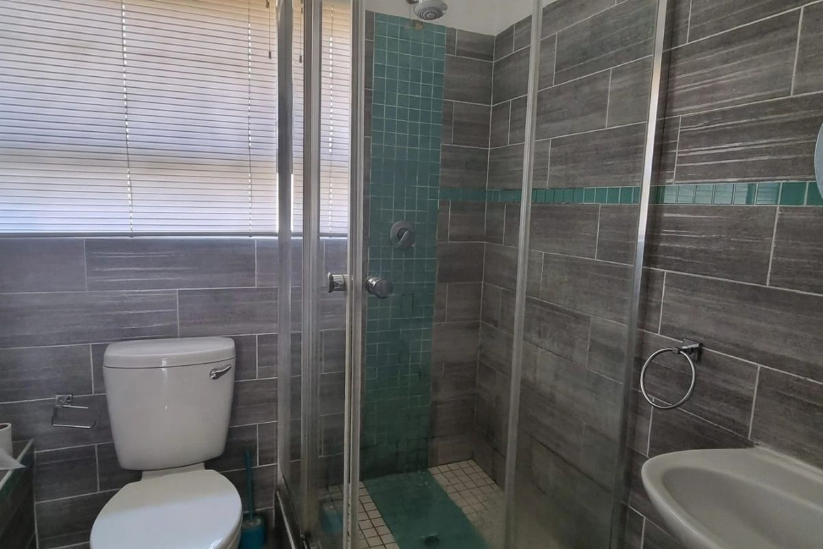 A functional bathroom featuring a glass shower enclosure, bathtub, pedestal basin, and grey tiled walls with teal accent detailing.