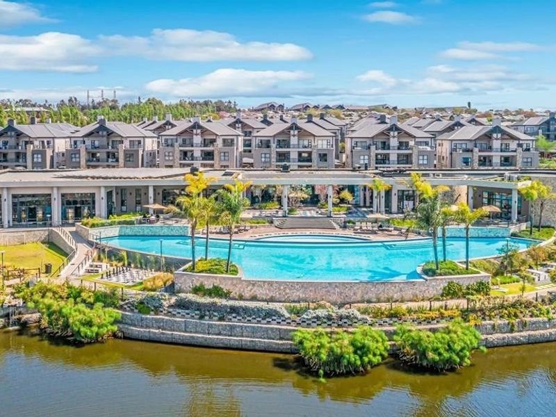 Aerial view of a large communal swimming pool and clubhouse facility set within a well-maintained residential apartment complex overlooking a lake.