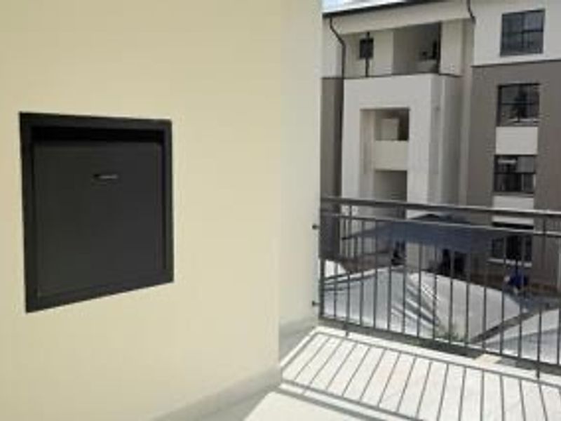 A clean, modern apartment balcony featuring a metal balustrade and a built-in wall safe, overlooking the complex courtyard.