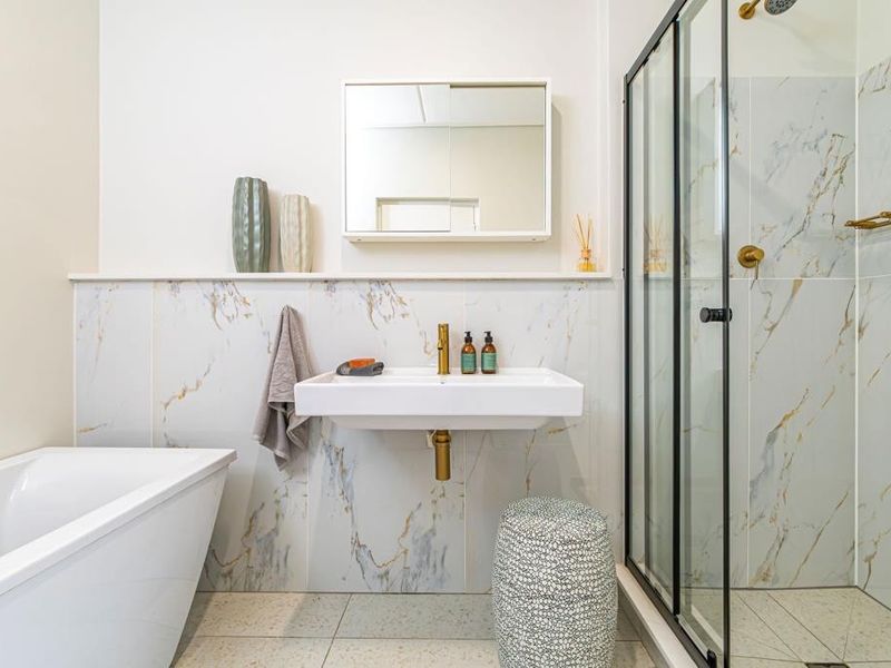 Modern bathroom featuring a freestanding bathtub, separate shower enclosure with black frame, wall-mounted basin, and elegant marble-look wall tiling with brass accents.