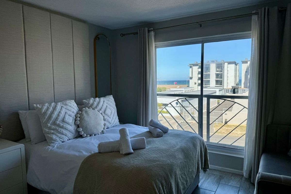 A well-maintained bedroom featuring floor-to-ceiling upholstered wall paneling, tiled flooring, and a sliding glass door opening to a balcony with coastal views.